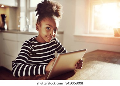 Thoughtful young black girl sitting watching the camera with a pensive expression as she browses the internet on a tablet computer at home