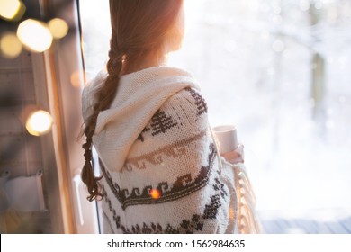 Thoughtful Young Brunette Woman Wearing Nordic Print Poncho Looking Through The Window, Blurry Winter Woods Snow Landscape Outside