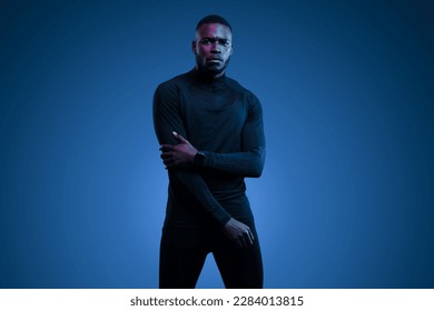 Thoughtful young brutal African American male model with dark hair and beard in total black activewear, with partial crossed arms looking at camera against blue background - Powered by Shutterstock