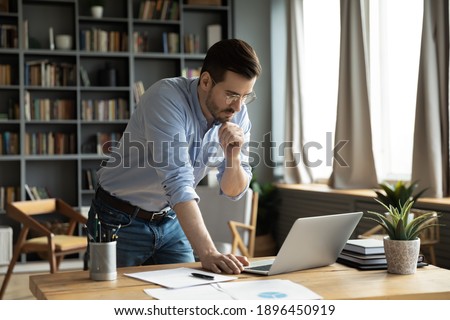 Thoughtful young businessman in eyeglasses working online on computer standing at table, analyzing report, making difficult decisions, developing company growth strategy at modern home office.