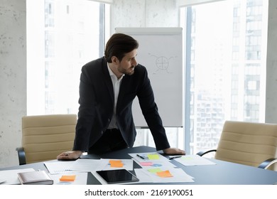Thoughtful young businessman in suit working in modern boardroom, stand in office lean at desk with papers, distracted from sales stats review, look aside, consider results. Workflow, business concept - Shutterstock ID 2169190051