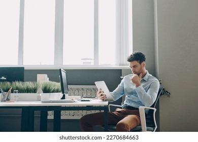 Thoughtful young businessman using digital tablet while sitting at his working place in office - Powered by Shutterstock