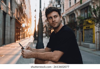 Hombre caucásico joven pensativo en camiseta negra con auriculares inalámbricos sentado en la calle de la ciudad durante el amanecer, sosteniendo el teléfono inteligente y mirando a la distancia, combinando tecnología con atención plena