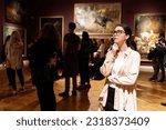 Thoughtful young Caucasian woman wearing glasses and looking at exhibition. In background, people are looking at paintings. Concept of Museum Day