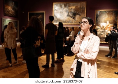 Thoughtful young Caucasian woman wearing glasses and looking at exhibition. In background, people are looking at paintings. Concept of Museum Day