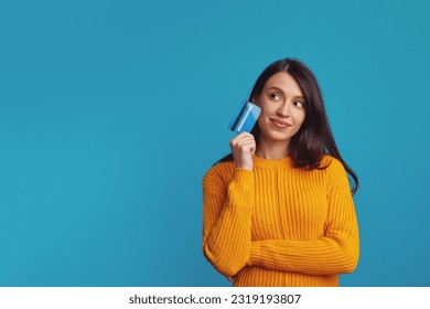 Thoughtful young girl wearing yellow clothes holding plastic credit card near face and looking at free empty space, isolated over blue background  - Powered by Shutterstock