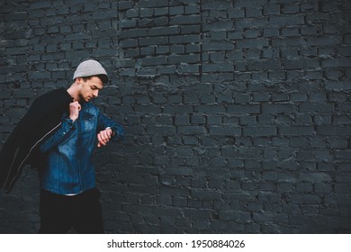 Thoughtful young hipster in denim shirt hat and black jacket on shoulder walking along dark brick wall checking time on wrist watch