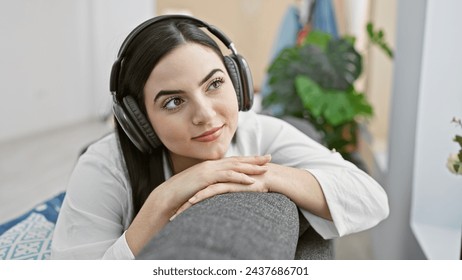 A thoughtful young hispanic woman with headphones relaxing at home on a couch near a houseplant - Powered by Shutterstock