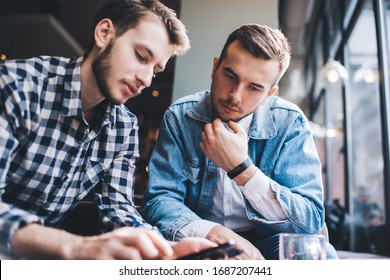 Thoughtful young male friends in  stylish casual clothing having conversation about application on mobile phone while sitting in cafe