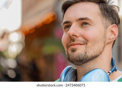 Thoughtful young male tourist in casual clothes with wireless headphones around neck looking up and smiling. Happy positive Caucasian man traveler standing on city urban street. Town lifestyles. - Powered by Shutterstock