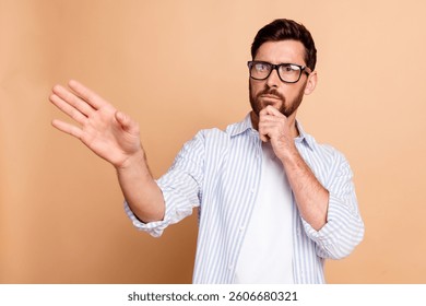 Thoughtful young man in casual striped shirt gesturing and pondering with thoughtful expression isolated against beige background