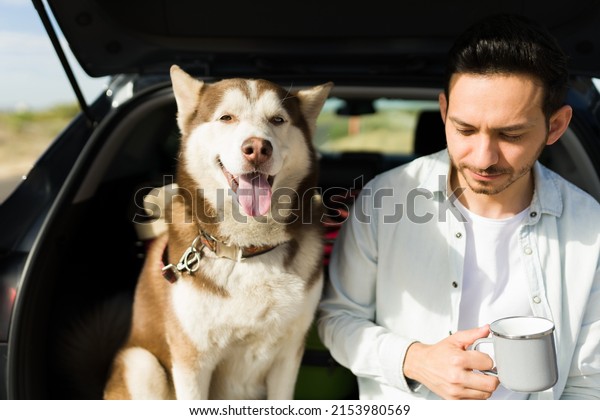 Thoughtful young man drinking\
coffee and resting while taking a vacation trip by car with his\
husky dog