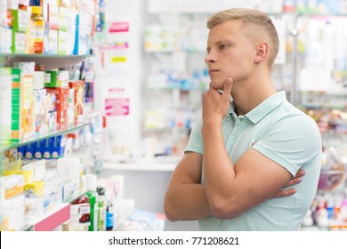 Thoughtful young man examining display at the local pharmacy shopping for prescripted medications at the drugstore looking thoughtful rubbing his chin doubting thinking medicine pharmaceutical.