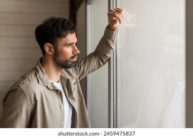 A thoughtful young man gazes out of a clear glass window, his hand gently resting on the pane, with sunlight softly illuminating his casual outfit and casting a glow on his contemplative face. - Powered by Shutterstock