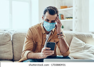 Thoughtful Young Man In Protective Face Mask Using Smart Phone While Spending Time At Home