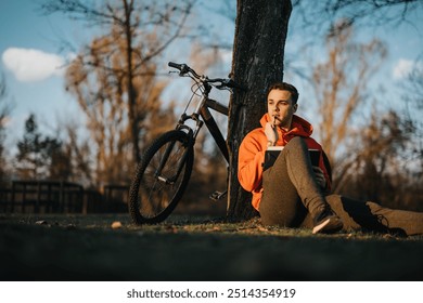 A thoughtful young man rests near his bike in a tranquil park setting as the sun sets, exuding a sense of leisure and freedom. - Powered by Shutterstock