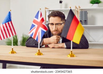 Thoughtful young man sitting at desk in office looking at USA, Great Britain and Germany flags. Businessman choosing foreign language or country to work or study in. Education or emigration - Shutterstock ID 2345427365