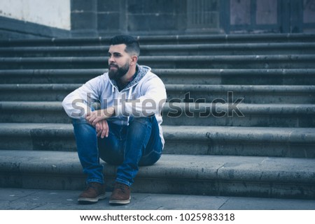 Thoughtful young man sitting outside church entrance stairs. Stylish male model with grey hoodie and denim jeans  alone on the street. Cold blue tones in urban city
