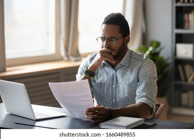 Thoughtful young mixed race businessman in glasses holding paper document, reading banking debt notification. Pensive millennial african american accountant employee reviewing research report. - Powered by Shutterstock