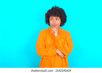 Thoughtful Young Sneaky Woman With Afro Hairstyle Wearing Orange Hoodie Against Blue Background Holds Chin And Looks Away Pensively Makes Up Great Plan