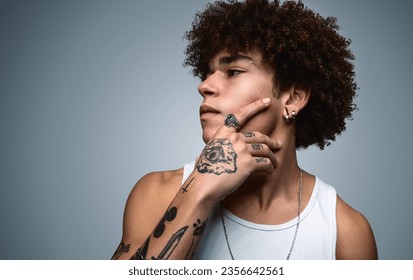 Thoughtful young tattooed Hispanic male model with dark curly hair in white top touching chin and looking away against gray background - Powered by Shutterstock