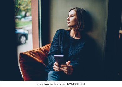 Thoughtful young woman dressed in black casual outfit looking out of window resting in comfortable coworking space. Pondering hipster girl with smartphone in hands thinking on future