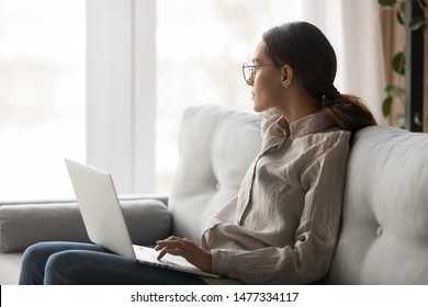 Thoughtful young woman in eyeglasses sitting with computer on couch, looking outside, cannot concentrate on work, need some rest, feeling bored, need additional motivation, working remotely at home.