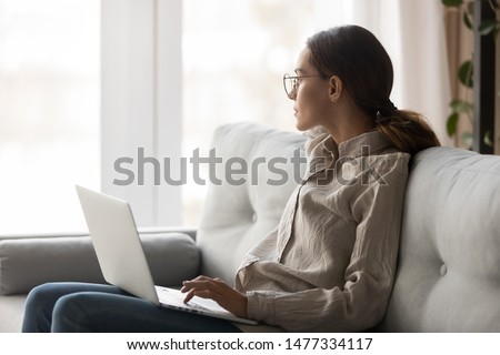 Thoughtful young woman in eyeglasses sitting with computer on couch, looking outside, cannot concentrate on work, need some rest, feeling bored, need additional motivation, working remotely at home.