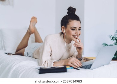 Thoughtful young woman with hair knot relaxing on bed with black purse browsing laptop touching lips and smiling in white room - Powered by Shutterstock