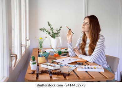 Thoughtful young woman holding paintbrush while painting on table at home - Powered by Shutterstock