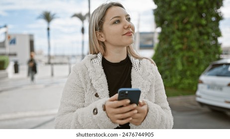 A thoughtful young woman holding a smartphone gazes away on a city street lined with palm trees. - Powered by Shutterstock