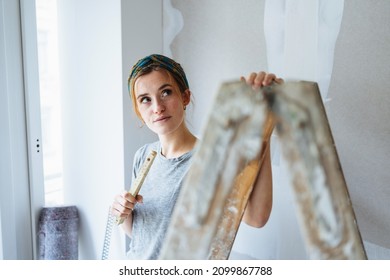 Thoughtful young woman inspecting her walls at home as she pauses during renovations and paint roller slung over her shoulder