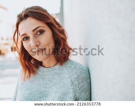 Thoughtful young woman scrutinising the camera with a pensive smile as she leans against a receding wall in a quiet city street