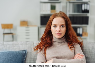 Thoughtful young woman staring intently ahead at the camera with a serious expression and folded arms as she relaxes on a sofa at home - Powered by Shutterstock