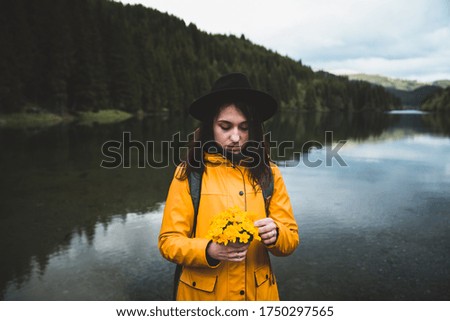 Thoughtful young woman wearing a hat, backpack and yellow jacket holding wild flowers bucket with lake and forest in the background. Travel and wanderlust concept. Girl is looking contemplative