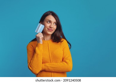 Thoughtful young woman wearing yellow clothes holding plastic credit card near face and looking at free empty space, isolated over blue background  - Powered by Shutterstock