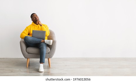 Thougthful African American Guy With Laptop Thinking Looking Aside Writing Article Working Online Sitting In Chair Over Gray Wall Background. Successful Freelance Career. Panorama, Copy Space
