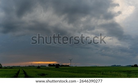 a threatening dark rain cloud over an agricultural field and wind turbines in East Friesland during sunset