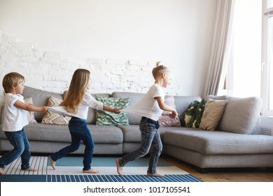 Three Active Barefooted Caucasian Kids Wearing Similar Clothes Celebrating Funny Birthday Party In Living Room, Dancing, Standing In Conga Line One After Another, Playing Indoor Games, Feeling Happy