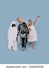 Three Adorable Little Kids In Halloween Costumes Over Blue Background. Ghost, Skeleton And Dead Bride Are Holding Hands. They Look Up, Pointing At Something.