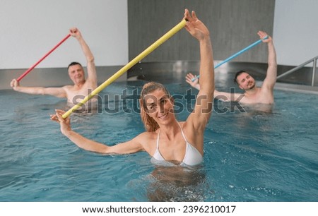 Three adults enjoy a water aerobics class, each holding different colored resistance bars aloft. Sports and gymnastics under water in swimming pool or spa resort.
