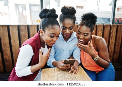 Three african american girls sitting on the table of caffe and looking on mobile phone.