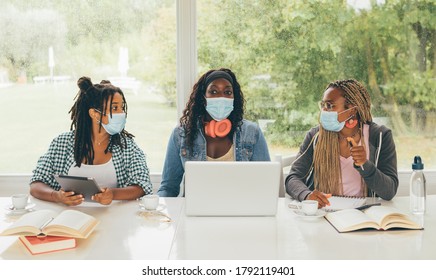 Three African American girls (students) sitting at the table studying up for test or making homework together, they are using laptop and digital tablet, wearing surgical masks. Back to school concept.