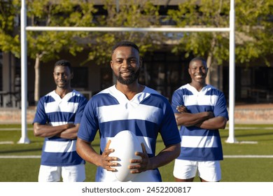 Three African American young male athletes holding a rugby ball, standing on field outdoors. All wearing striped jerseys, shorts, and have short hair, unaltered. - Powered by Shutterstock