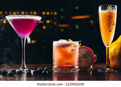 Three Alcoholic Cocktails, Purple, Orange And Yellow Colors On The Table. Variety Of Colorful Alcoholic Drinks 