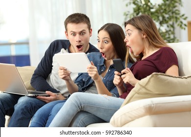 Three Amazed Friends On Line With Multiple Devices Sitting On A Sofa In The Living Room In A House Interior