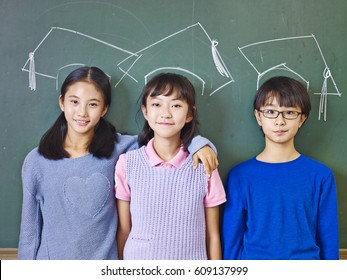 Three Asian Elementary School Children Standing In Front Of Chalkboard Underneath Chalk-drawn Graduation Caps.