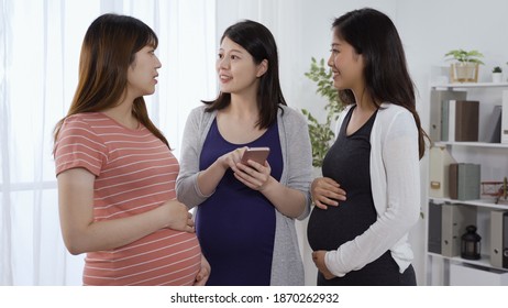 Three Asian Maternity Moms With Big Belly Standing Together And Looking At Information About Prenatal Classes Online On Mobile Phone.