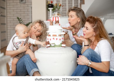 Three Attractive Middle-aged And Young Women Blowing Kisses To Cute Little One-year-old Boy Touching Bear On Birthday Cake With Macaroons Celebrating At Home. Family, Celebration, Birthday Concept.