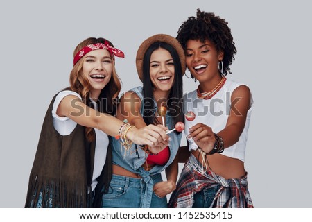 Three attractive young women eating lollipop and smiling while standing against grey background